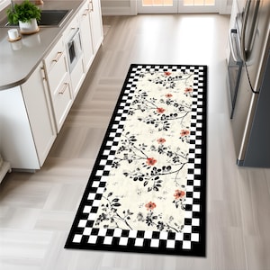 Puede incluir: Una alfombra de cocina rectangular con un diseño floral. La alfombra presenta un centro color crema con ramas negras, flores rojas y hojas verdes. Un borde a cuadros blanco y negro enmarca el patrón floral.