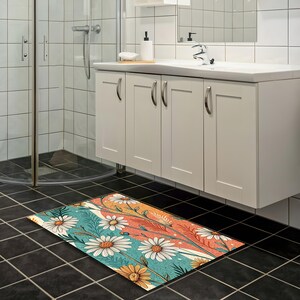 Op de afbeelding: Een rechthoekige badmat met een bloemenontwerp met grote witte madeliefjes, gele bloemen en groene bladeren. De achtergrond heeft strepen in turquoise, oranje en crème kleuren. De mat ligt op een zwarte tegelvloer.