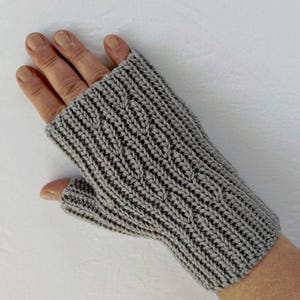 Op de afbeelding: Een grijze gebreide vingerloze handschoen met een kabelpatroon. De handschoen is gemaakt van zacht, getextureerd garen en heeft een geribbelde manchet. De handschoen is ontworpen om de handen warm te houden terwijl de vingers vrij blijven. Ongeveer 20 cm lang.