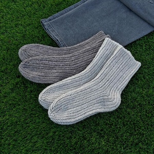 May include: Two pairs of knitted socks in shades of gray, one pair a darker charcoal and the other a lighter heather gray. The socks are ribbed and appear to be made of a soft, warm material. A pair of blue jeans is folded in the background.