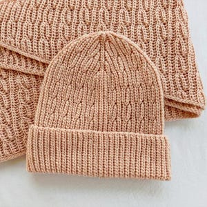 Puede incluir: Un conjunto de gorro y bufanda de punto en color marrón claro. El gorro tiene un borde doblado y un patrón texturizado. La bufanda está doblada y muestra un patrón a juego. El conjunto está hecho de un material suave y cálido.