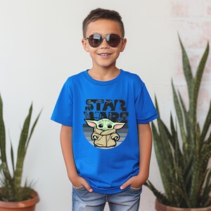 以下が含まれることがあります： スターウォーズのグローグのグラフィックが特徴の青いTシャツ。デザインには、グローグの上に「STAR WARS」という言葉が含まれています。サングラスをかけた子供がTシャツを着ています。