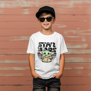 以下が含まれることがあります： スターウォーズのグローグーのグラフィックと「STAR WARS」の文字が入った白いTシャツ。黒いキャップとサングラスをかけた子供が着ています。