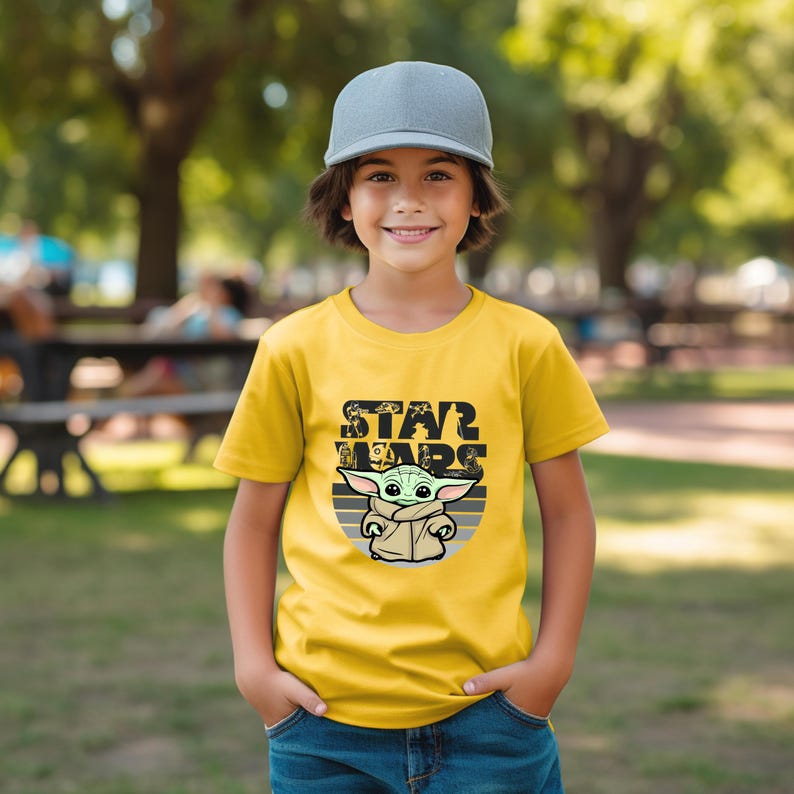 以下が含まれることがあります： スターウォーズのグローグのグラフィックと「STAR WARS」の文字が入った黄色のTシャツ。子供はグレーの野球帽をかぶっています。画像は屋外で撮影されました。
