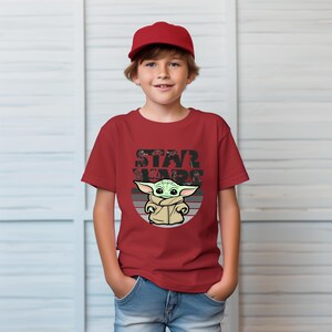 以下が含まれることがあります： スターウォーズのグローグのグラフィックが特徴の赤いTシャツ。 「STAR WARS」の文字がグローグの画像の上にあります。 子供は赤い野球帽をかぶっています。