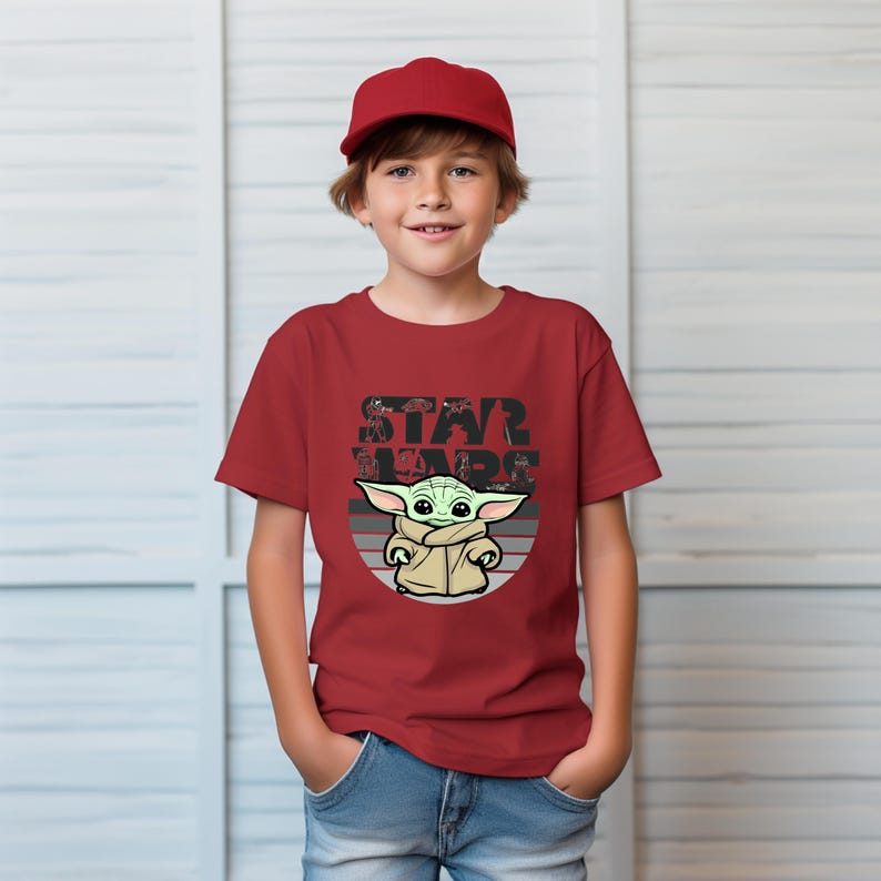 以下が含まれることがあります： スターウォーズのグローグのグラフィックが特徴の赤いTシャツ。 「STAR WARS」の文字がグローグの画像の上にあります。 子供は赤い野球帽をかぶっています。