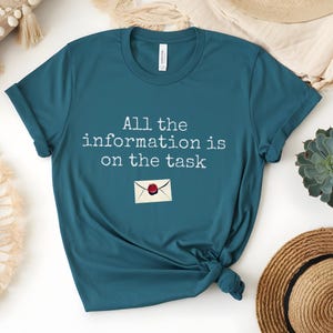 May include: Teal t-shirt with the text "All the information is on the task" and an envelope graphic. The shirt is knotted at the bottom. A succulent, a straw hat, and a decorative pillow are in the background.