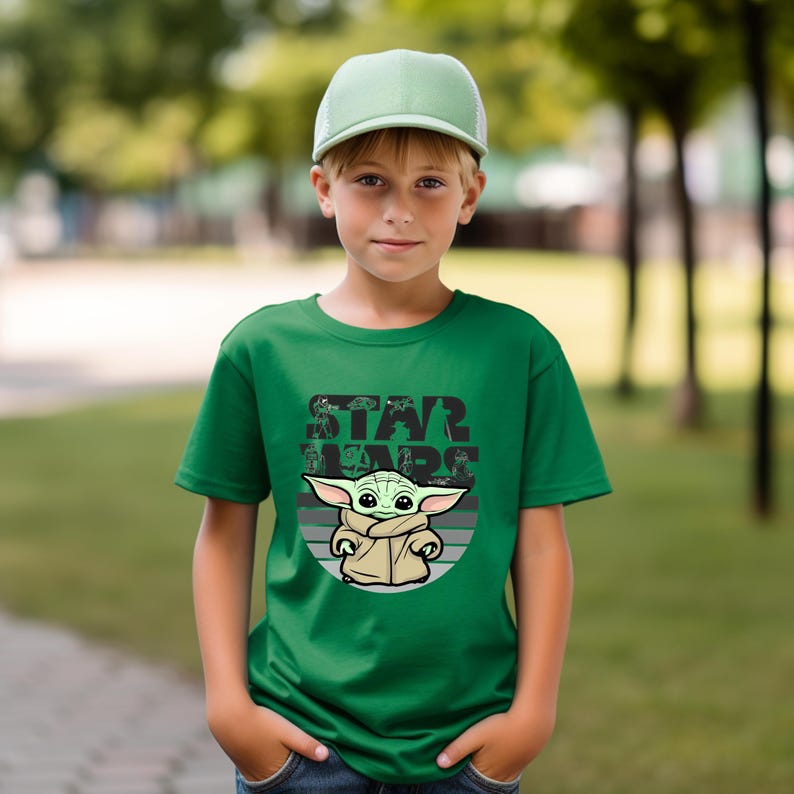 以下が含まれることがあります： スターウォーズのベビーヨーダのグラフィックと「STAR WARS」の文字が入った緑色のTシャツ。写真の人物は緑色の野球帽をかぶっています。背景は緑の木々でぼやけています。