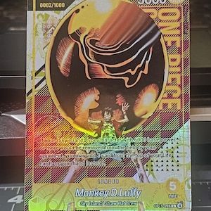 May include: A holographic trading card featuring Monkey D. Luffy from the Sky Island/Straw Hat Crew. The card has a gold and yellow color scheme with the number 5000 and the text "ONE PIECE" visible. The card also includes the text "Monkey.D.Luffy".