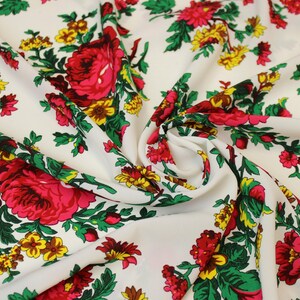 Op de afbeelding: Witte stof met een levendig bloemenpatroon. Grote rode en roze bloemen worden afgewisseld met kleinere gele bloesems en groene bladeren. De stof is opgevouwen, wat een dynamische weergave van het ontwerp creëert.