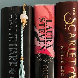 May include: A bookmark featuring a light blue braided cord and a matching tassel, accented with a gold-toned metal ring and a white, textured jellyfish charm with gold-toned tentacles, positioned between three hardcover books on a shelf.
