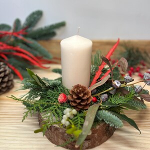 May include: A Christmas-themed centrepiece with a cream pillar candle, greenery, red berries, and a pine cone, set in a rustic wooden base. Ideal for festive home decoration.
