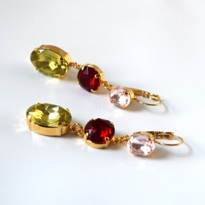 May include: A pair of gold earrings with three oval-shaped gemstones. The top gemstone is light green, the middle gemstone is red, and the bottom gemstone is pink.