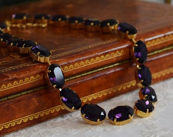 May include: A purple gemstone necklace with oval-shaped stones set in gold. The necklace is lying on a brown leather surface.