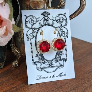 May include: A pair of red gemstone earrings with gold settings. The earrings are displayed on a white card with the text "Dames a la Mode".