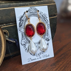 May include: Gold-toned earrings with a red, oval-shaped gemstone and a white teardrop-shaped pearl. The earrings are displayed on a card with the text "Dames a la Mode". The background includes a vintage green box.