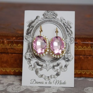 Puede incluir: Un par de pendientes ovalados de color rosa con engastes dorados y un borde de strass transparente. Los pendientes se muestran en una tarjeta blanca con el texto "Dames a la Mode".