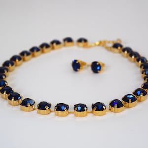 May include: A gold necklace with a row of dark blue rhinestones and matching earrings. The necklace is made of small, round rhinestones that are connected by a gold chain.