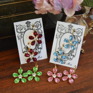 May include: Four pairs of flower-shaped earrings in various colors: red, blue, green, and pink. Each earring features a central crystal surrounded by petals. The earrings are displayed on white cards with decorative borders.