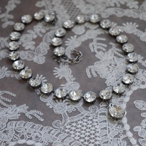 May include: A silver necklace with a teardrop pendant, made of clear faceted crystals. The necklace is on a white lace background.