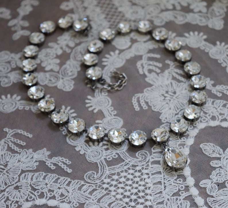 May include: A silver necklace with a teardrop pendant, made of clear faceted crystals. The necklace is on a white lace background.