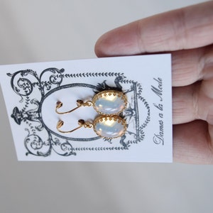 Pode incluir: Um par de brincos de ouro com pedras opalescentes de forma oval. Os brincos t&ecirc;m um delicado design de filigrana dourada em torno das pedras. Os brincos s&atilde;o exibidos em um cart&atilde;o branco com o texto "Dames a la Mode".