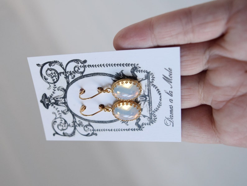 Pode incluir: Um par de brincos de ouro com pedras opalescentes de forma oval. Os brincos t&ecirc;m um delicado design de filigrana dourada em torno das pedras. Os brincos s&atilde;o exibidos em um cart&atilde;o branco com o texto "Dames a la Mode".