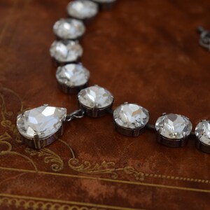 May include: A silver necklace with a teardrop-shaped pendant and round crystal stones. The necklace is on a brown leather surface.