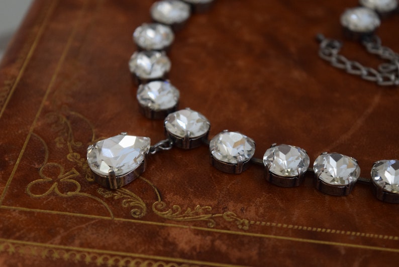 May include: A silver necklace with a teardrop-shaped pendant and round crystal stones. The necklace is on a brown leather surface.