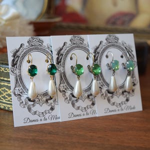 May include: Three pairs of dangle earrings displayed on white cards. Each earring features a gold-colored hook, a green gemstone, and a white teardrop-shaped pearl. The cards have a decorative black and white design and the text "Dames a la Mode".