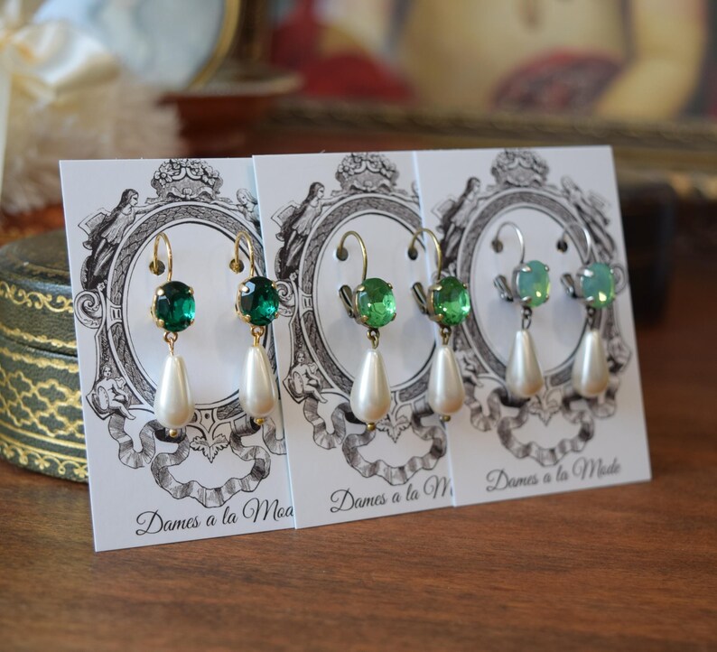 May include: Three pairs of dangle earrings displayed on white cards. Each earring features a gold-colored hook, a green gemstone, and a white teardrop-shaped pearl. The cards have a decorative black and white design and the text "Dames a la Mode".