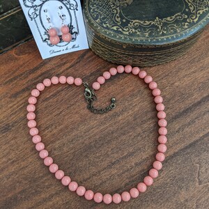 以下が含まれることがあります： 銀色の留め具が付いたピンクのビーズネックレス。ネックレスは木製の表面の上に置かれています。ネックレスには、「Dames a la Mode」と書かれたカードにピンクのイヤリングが1組付いています。