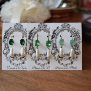 May include: Three pairs of elegant earrings displayed on cards. Each earring features a green gemstone and a dangling white teardrop-shaped pearl. The earrings have gold-colored hooks. The cards have a vintage design with the text "Dames a la Mode".