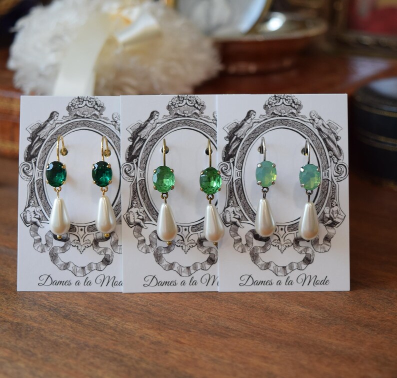 May include: Three pairs of elegant earrings displayed on cards. Each earring features a green gemstone and a dangling white teardrop-shaped pearl. The earrings have gold-colored hooks. The cards have a vintage design with the text "Dames a la Mode".