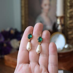 May include: Gold-toned earrings featuring oval emerald green gemstones and teardrop-shaped faux pearls. The earrings have a classic design with a lever-back closure. The gemstones are set in a gold bezel, and the pearls are a creamy white color.