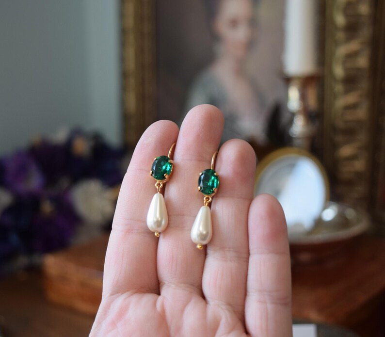 May include: Gold-toned earrings featuring oval emerald green gemstones and teardrop-shaped faux pearls. The earrings have a classic design with a lever-back closure. The gemstones are set in a gold bezel, and the pearls are a creamy white color.