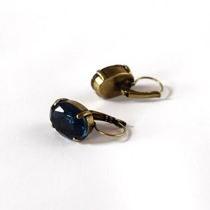 Puede incluir: Un par de pendientes de estilo antiguo con piedras preciosas ovaladas, facetadas, de color azul oscuro. Los pendientes tienen un engaste de metal color bronce y cierres de palanca. Las piedras preciosas son el punto focal, a&ntilde;adiendo elegancia vintage.