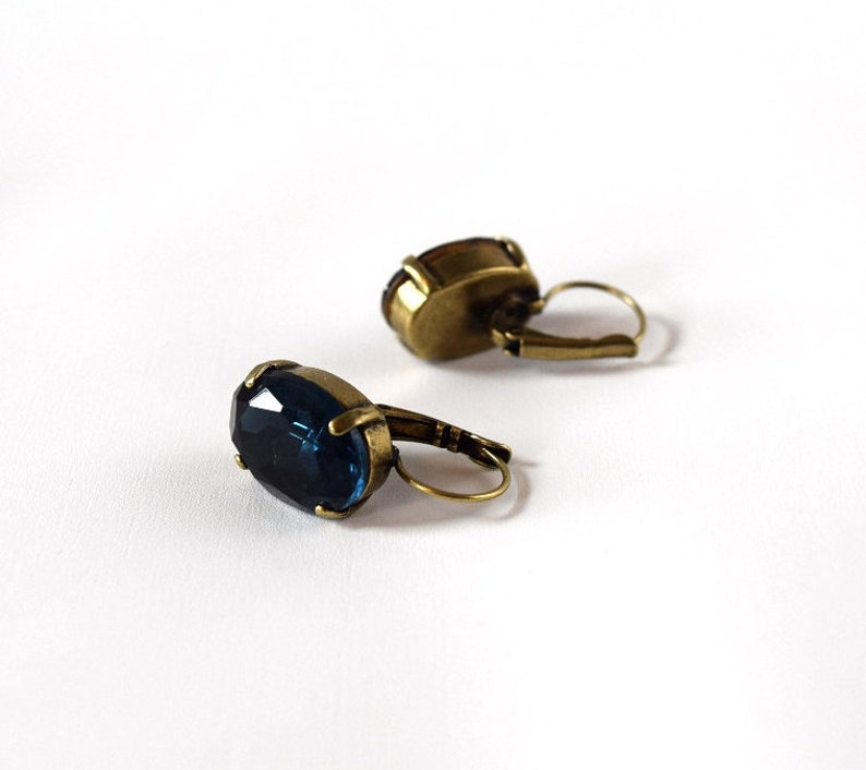 Puede incluir: Un par de pendientes de estilo antiguo con piedras preciosas ovaladas, facetadas, de color azul oscuro. Los pendientes tienen un engaste de metal color bronce y cierres de palanca. Las piedras preciosas son el punto focal, a&ntilde;adiendo elegancia vintage.