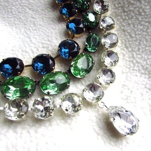 May include: A multi-strand necklace with large oval-shaped rhinestones in shades of green, blue, and clear. The necklace has a teardrop-shaped pendant at the end.