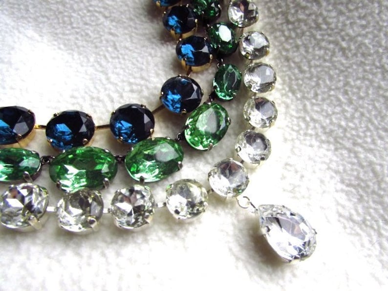 May include: A multi-strand necklace with large oval-shaped rhinestones in shades of green, blue, and clear. The necklace has a teardrop-shaped pendant at the end.