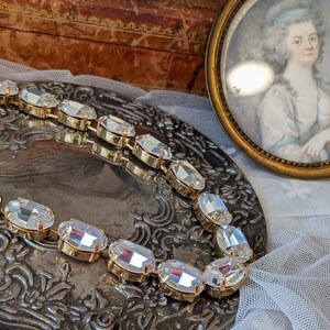 Puede incluir: Un collar de tono dorado que presenta una serie de cristales transparentes de forma ovalada y tallados, montados en engastes met&aacute;licos individuales. El collar est&aacute; dispuesto en una l&iacute;nea curva, resaltando el brillo de las piedras y el acabado met&aacute;lico.