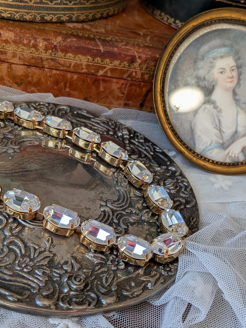 Puede incluir: Un collar de tono dorado que presenta una serie de cristales transparentes de forma ovalada y tallados, montados en engastes met&aacute;licos individuales. El collar est&aacute; dispuesto en una l&iacute;nea curva, resaltando el brillo de las piedras y el acabado met&aacute;lico.