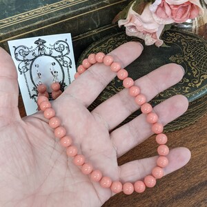 以下が含まれることがあります： 珊瑚ピンクのビーズネックレスと、それに合わせたイヤリングのセット。ネックレスは丸いビーズで作られており、長さは約45cmです。