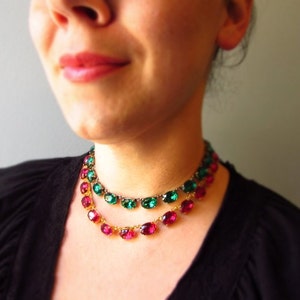 Puede incluir: Un collar de dos hileras con piedras ovaladas facetadas. La hilera superior presenta piedras verde esmeralda engastadas en metal, mientras que la inferior muestra piedras de color rosa magenta en una cadena dorada.