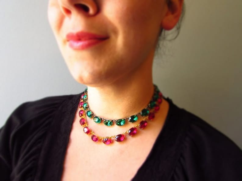 Puede incluir: Un collar de dos hileras con piedras ovaladas facetadas. La hilera superior presenta piedras verde esmeralda engastadas en metal, mientras que la inferior muestra piedras de color rosa magenta en una cadena dorada.
