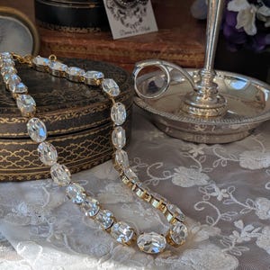 Puede incluir: Un collar de tono dorado que presenta una fila continua de cristales transparentes de forma ovalada y facetada, engastados en biseles de metal. El collar est&aacute; dispuesto en una l&iacute;nea curva, mostrando el brillo de las piedras y los eslabones met&aacute;licos.