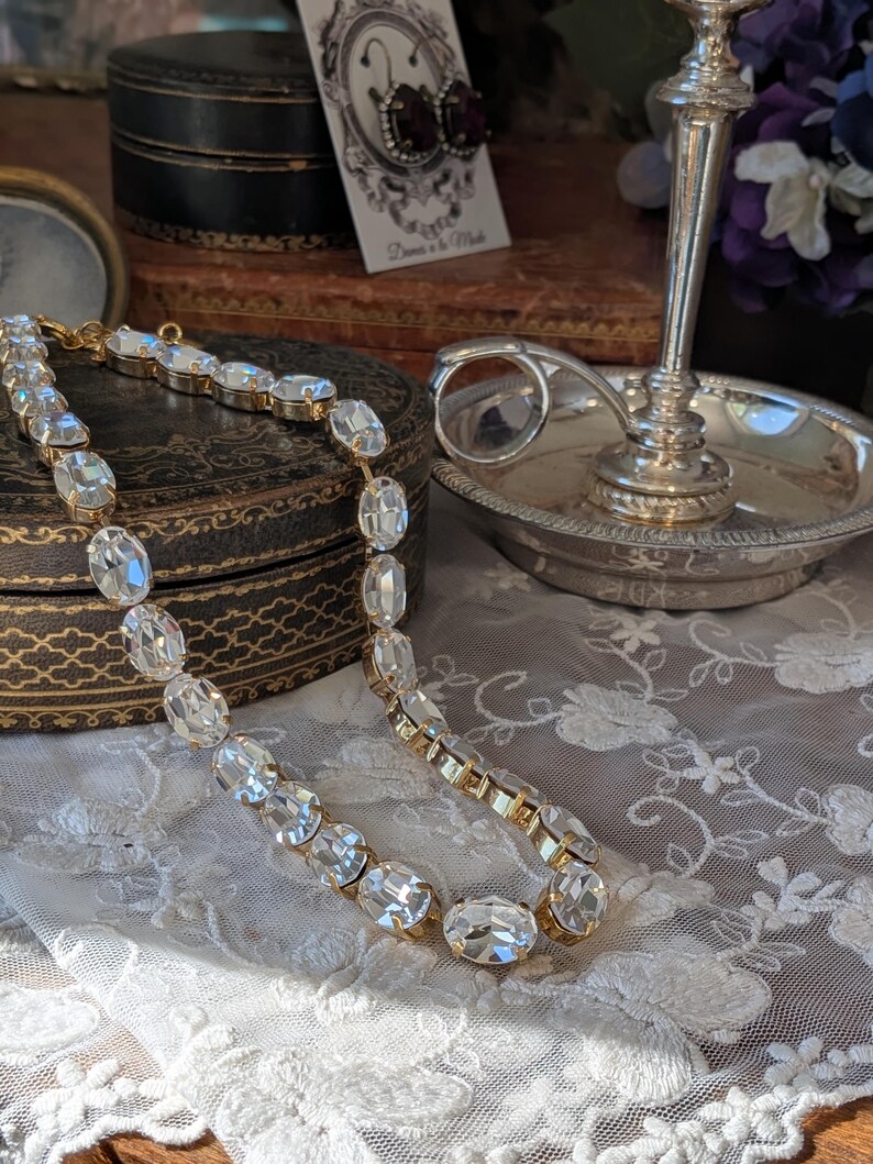 Puede incluir: Un collar de tono dorado que presenta una fila continua de cristales transparentes de forma ovalada y facetada, engastados en biseles de metal. El collar est&aacute; dispuesto en una l&iacute;nea curva, mostrando el brillo de las piedras y los eslabones met&aacute;licos.