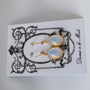Pode incluir: Um par de brincos de ouro com pedras de opala branca. Os brincos est&atilde;o em um cart&atilde;o branco com o texto "Dames a la Mode".