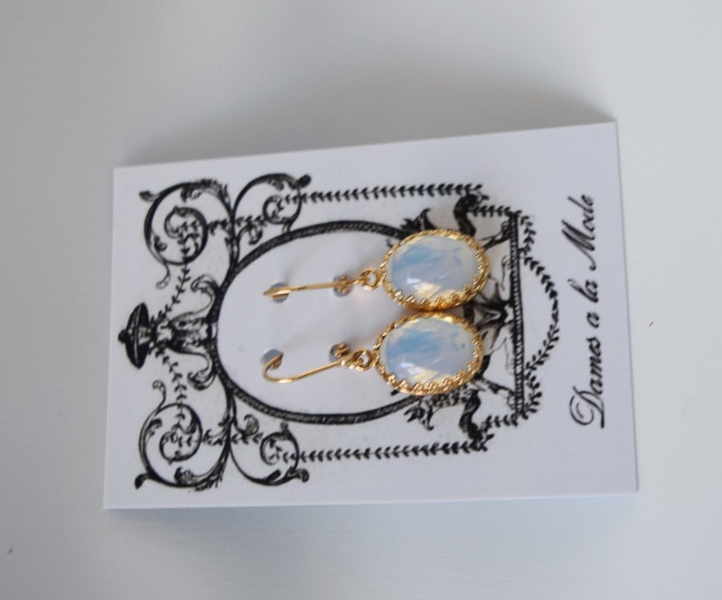 Pode incluir: Um par de brincos de ouro com pedras de opala branca. Os brincos est&atilde;o em um cart&atilde;o branco com o texto "Dames a la Mode".