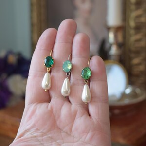 May include: Three pairs of elegant drop earrings. Each earring features a gold-colored hook, a green gemstone, and a pear-shaped faux pearl. The earrings are displayed against a soft, blurred background, highlighting their delicate design.